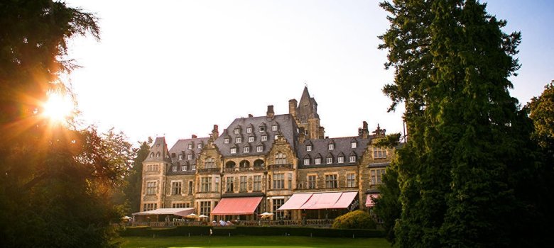 Eine Außenansicht des historischen Schlosshotels Kronberg inmitten einer gepflegten Parkanlage im warmen Gegenlicht der tiefstehenden Sonne.