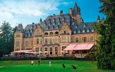 Mehrere Personen beim Golfspielen auf einem grünen Rasen vor der detailreichen, von Bäumen umgebenen Steinfassade des historischen Schlosshotels Kronberg.