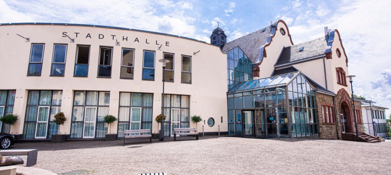 Weitwinkelaufnahme des gesamten Gebäudekomplexes unter blauem Himmel mit lockeren Wolken. Zu sehen ist die moderne, helle Fassade der Stadthalle (links), ein zentraler Glaseingang und ein angrenzendes historisches Gebäude aus Naturstein mit markantem Giebel (rechts)