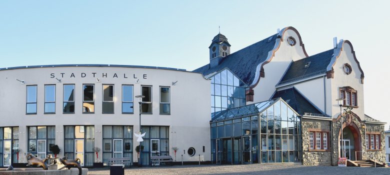 Ansicht der Stadthalle in klarem Winterlicht. Im linken Vordergrund ist ein Brunnen mit bronzenen Tierfiguren (Delfine oder Robben) zu sehen. Das historische Gebäude auf der rechten Seite ist mit einem weihnachtlichen Stern geschmückt