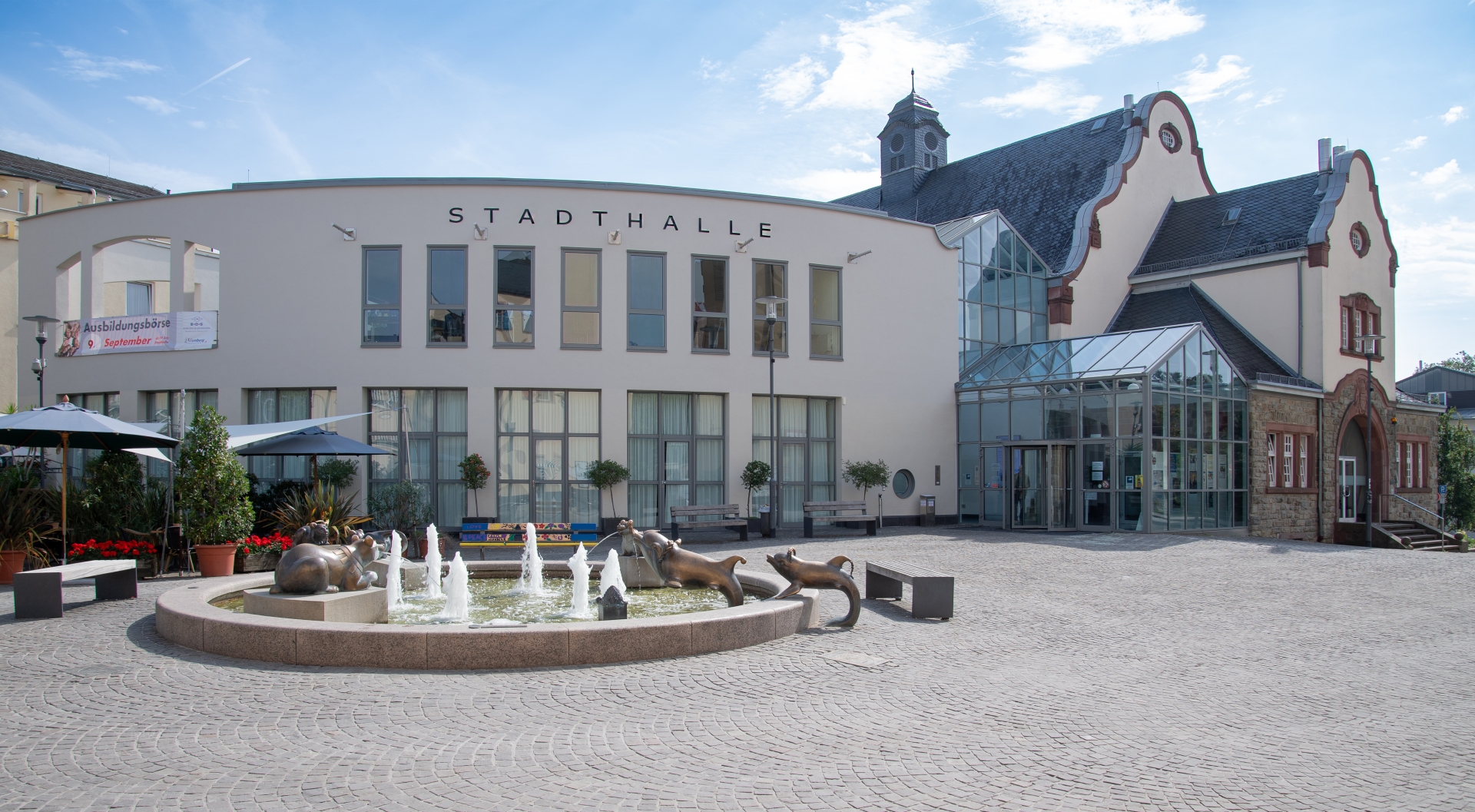 Zentrale Ansicht des Stadthallenvorplatzes mit Fokus auf den Springbrunnen und die Tierplastiken. Im Hintergrund ist an der modernen Fassade ein Banner für eine „Ausbildungsbörse“ zu erkennen. Der Platz ist weiträumig mit Kopfsteinpflaster gestaltet