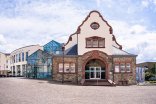 Nahaufnahme des historischen Gebäudeteils der Stadthalle. Das Gebäude aus Naturstein verfügt über einen großen, treppenversetzten Rundbogeneingang, ein Wappenrelief über der Tür und einen kunstvoll gestalteten Giebel