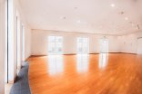 Ein großer, vollständig leerer Saal mit glänzendem Parkettboden, weißen Wänden und mehreren großen Fenstern sowie einer verglasten Tür an der Stirnseite.