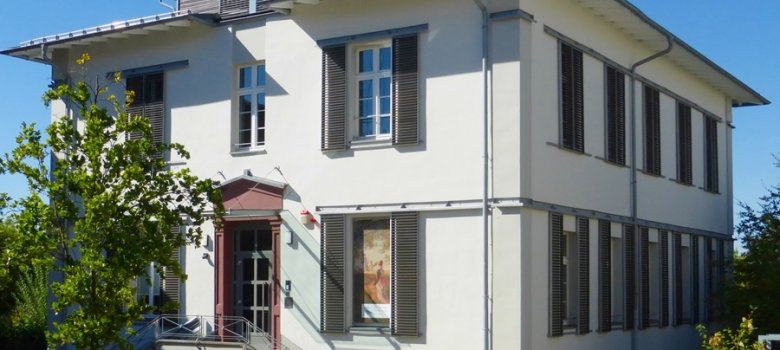 Ein zweistöckiges, helles Gebäude mit einer markanten Außentreppe, dunklen Fensterläden und einem modernen Dachaufbau unter strahlend blauem Himmel.