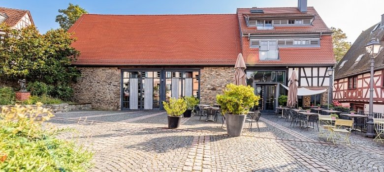 Ein sonniger, kopfsteingepflasterter Platz vor einem historischen Gebäude mit Natursteinmauer, Fachwerkelementen, einem roten Ziegeldach und einer bestuhlten Außengastronomie.