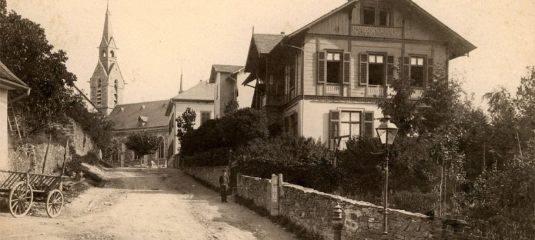 Historische Sepia-Fotografie einer unbefestigten Straße in Kronberg im Taunus, die bergauf an einer Natursteinmauer und Wohnhäusern vorbei zu einer Kirche mit spitzem Kirchturm führt.