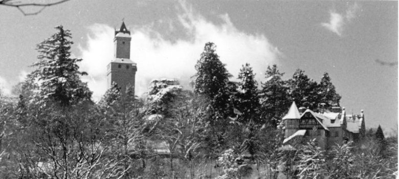 Historische Schwarz-Weiß-Fotografie einer verschneiten Winterlandschaft in Kronberg mit der Burg Kronberg, dem Freischütz-Heim auf einer Anhöhe und Menschen auf einer schneebedeckten Freifläche im Vordergrund.