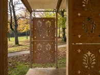 Fotografie des überdachten Quellenpavillons im Quellenpark Kronthal mit verzierten, durchbrochenen Holzwänden, die den Blick auf die umliegenden herbstlichen Bäume und Wiesen freigeben.