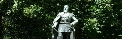 Fotografie des Kaiser-Friedrich-Denkmals in Kronberg im Taunus, das eine bronzene Standbild-Statue von Kaiser Friedrich III. auf einem verzierten Steinsockel vor dichtem, grünem Hintergrund aus Bäumen zeigt.