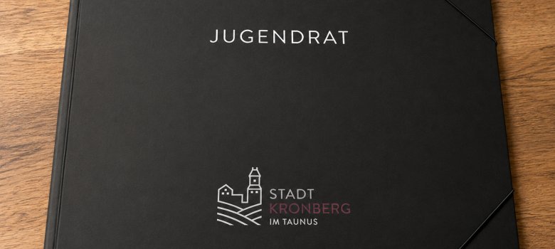 Jugendrat Mappe Eine geschlossene, schwarze Dokumentenmappe mit der Aufschrift „JUGENDRAT“ und dem Logo der Stadt Kronberg im Taunus auf einem Holztisch.
