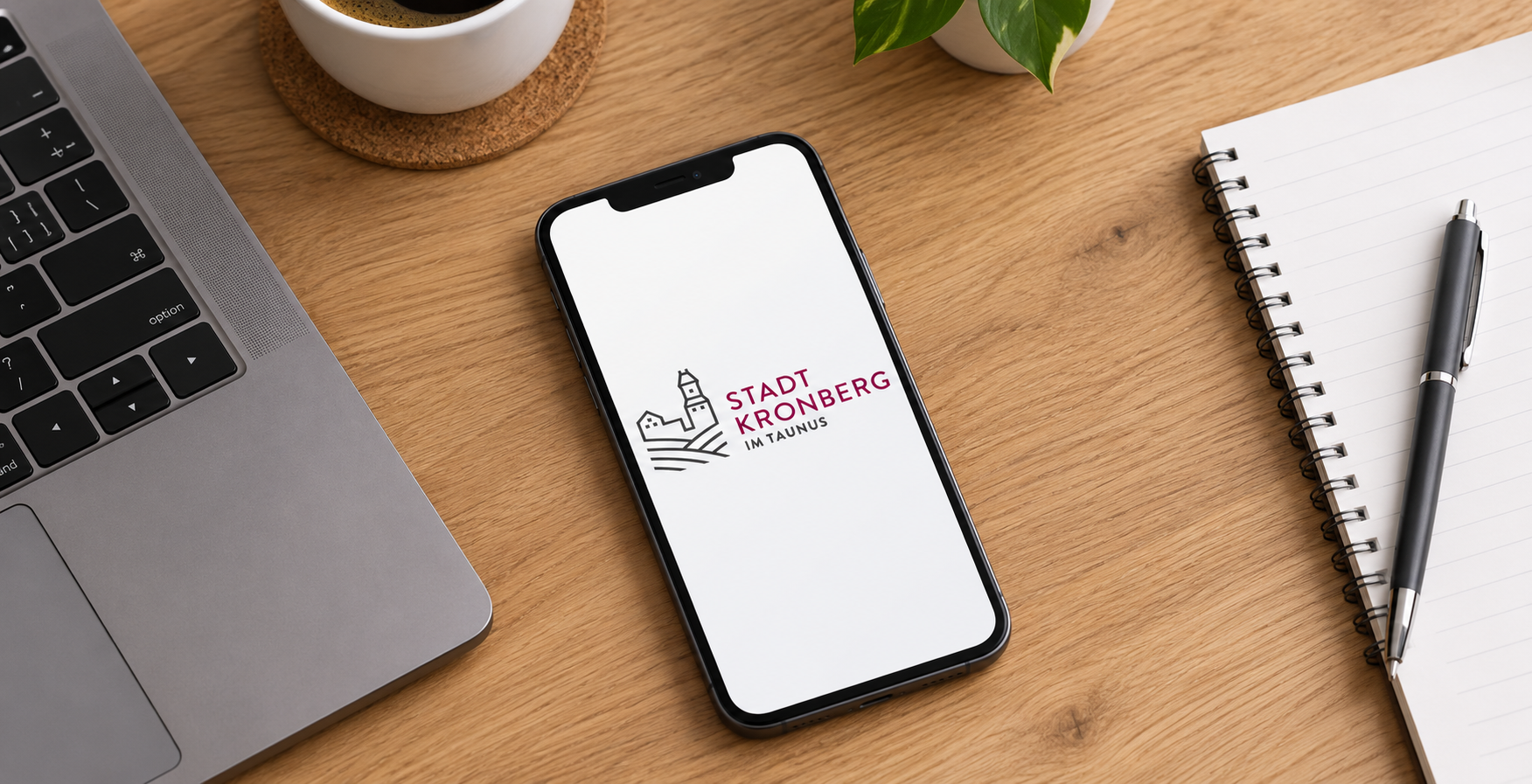 Minimalistischer Arbeitsplatz mit Stadtlogo Ein Smartphone liegt flach auf einem hellen Holztisch und zeigt auf dem Display das Logo der „Stadt Kronberg im Taunus“. Daneben befinden sich die Tastatur eines Laptops sowie ein geöffnetes Notizbuch mit einem Stift