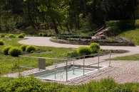Ein im Boden eingelassenes, rechteckiges Kneippbecken mit klarem Wasser, umgeben von einer gepflasterten Fläche und Edelstahlgeländern zum Festhalten. Die Anlage befindet sich in einer gepflegten Parklandschaft mit Rasenflächen, kugelig geschnittenen Sträuchern und einem geschwungenen Spazierweg