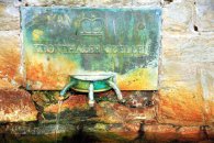 Nahaufnahme einer verwitterten, grünlich patinierten Metalltafel mit der Aufschrift „KRONTHALER QUELLEN“ und einem Kronen-Symbol. Unter der Tafel fließt aus einem von drei metallenen Rohren Wasser in ein steinernes Becken