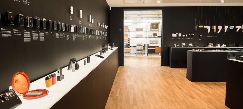 Eine moderne Museumsgalerie mit hellem Parkettboden und mattschwarzen Wänden. An der linken Wand sind zahlreiche historische Braun-Geräte, wie Rasierer und Taschenrechner, in chronologischen Reihen ausgestellt. Weiße Podeste präsentieren weitere Designobjekte. Ein Durchgang im Hintergrund gibt den Blick auf einen weiteren hellen Ausstellungsraum frei
