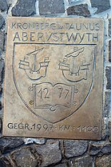 Eine rechteckige Bronzeschild-Plakette ist auf einer rustikalen Natursteinmauer angebracht. Sie zeigt oben die Schriftzüge „KRONBERG IM TAUNUS“ und darunter „ABERYSTWYTH“ sowie ein zentrales Wappen mit zwei Segelschiffen über einem aufgeschlagenen Buch, das die Zahl „12 77“ trägt. Am unteren Rand steht die Gravur „GEGR. 1997 - KM : 1100“