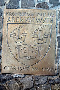 Eine rechteckige Bronzeschild-Plakette ist auf einer rustikalen Natursteinmauer angebracht. Sie zeigt oben die Schriftzüge „KRONBERG IM TAUNUS“ und darunter „ABERYSTWYTH“ sowie ein zentrales Wappen mit zwei Segelschiffen über einem aufgeschlagenen Buch, das die Zahl „12 77“ trägt. Am unteren Rand steht die Gravur „GEGR. 1997 - KM : 1100“