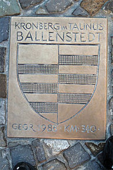 Eine quadratische Bronzeplakette ist bündig in ein dunkles Kopfsteinpflaster eingelassen. Sie trägt oben die eingravierten Namen der Partnerstädte „KRONBERG IM TAUNUS“ und „BALLENSTEDT“, darunter ein großes, geteiltes Wappenschild mit horizontalen Streifenmustern und unten die Inschrift „GEGR. 1988 - KM : 340