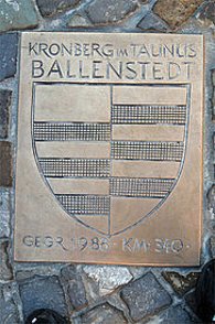 Eine quadratische Bronzeplakette ist bündig in ein dunkles Kopfsteinpflaster eingelassen. Sie trägt oben die eingravierten Namen der Partnerstädte „KRONBERG IM TAUNUS“ und „BALLENSTEDT“, darunter ein großes, geteiltes Wappenschild mit horizontalen Streifenmustern und unten die Inschrift „GEGR. 1988 - KM : 340
