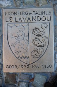 Eine hochkant rechteckige Bronzeplakette ist in Kopfsteinpflaster eingelassen und erinnert an die Städtepartnerschaft zwischen „KRONBERG IM TAUNUS“ und „LE LAVANDOU“. Das zentrale Wappenschild zeigt links einen aufrecht stehenden Löwen unter einer Krone und rechts drei untereinander angeordnete Delfine; am unteren Rand stehen die Gravur „GEGR. 1972“ und die Entfernungsangabe „KM: 1150“