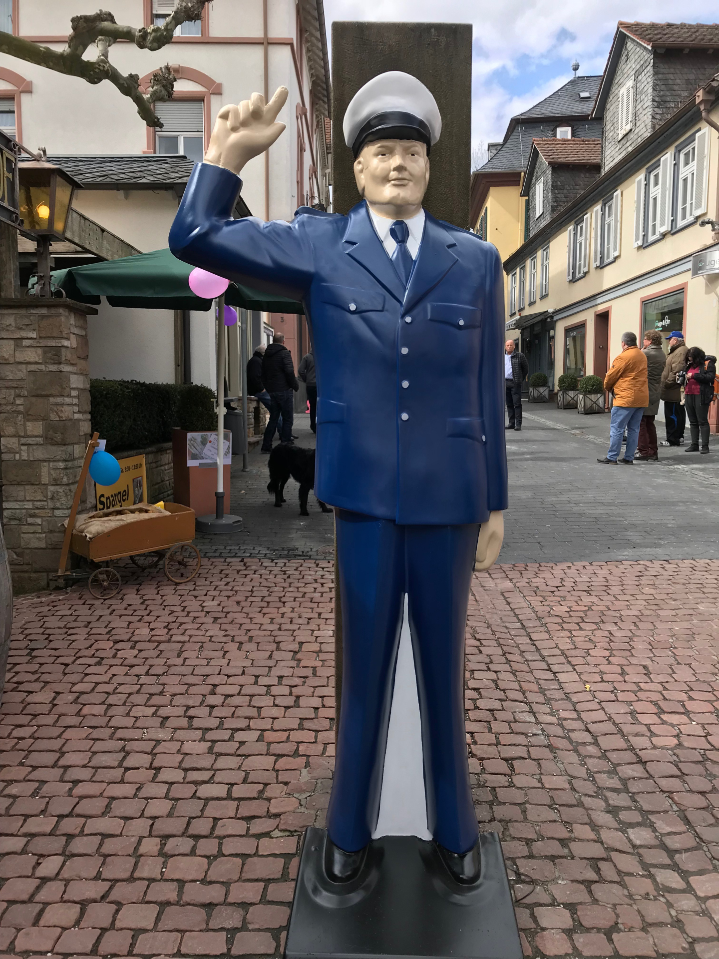 Eine lebensgroße, farbige Skulptur eines Polizisten in einer klassischen blauen Uniform mit Schirmmütze. Er steht auf Kopfsteinpflaster und hat den rechten Arm zum Gruß oder Zeichengeben erhoben. Im Hintergrund ist eine Fußgängerzone mit Passanten und Fachwerkhäusern zu sehen.