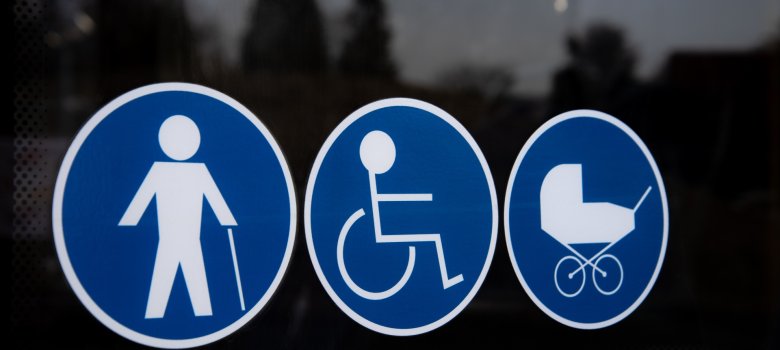 Drei blaue, kreisförmige Piktogramme auf einer Glasscheibe: Eine Person mit Gehstock, eine Person im Rollstuhl und ein Kinderwagen