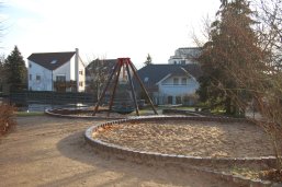 Ein Spielplatz mit einer runden Sandkiste und einem Klettergerüst vor Wohnhäusern