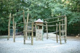 Ein rustikales Spielgerät aus Naturholz mit Kletternetzen, Seilbrücken und einem kleinen Holzturm auf einer hellen Kiesfläche vor dichtem Waldhintergrund