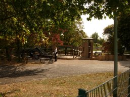 Ein sonniger Spielplatz mit einem hölzernen Spielturm, einer Rutsche und Sitzbänken, umgeben von herbstlich gefärbten Bäumen und einem grünen Zaun im Vordergrund