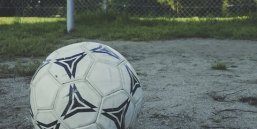 Fußball, der auf sandigen Grund vor einer Wiese mit einem Zaun liegt