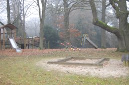 Links im Bild ein Holztürmchen mit Rutsche. in der Mitte ein Sandkasten und im Hintergrund 2 Wippen sowie eine weitere Rutsche. Es stehen mehrere Bäume verteilt um und auf dem Spielplatz