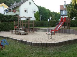 Sandkasten, in den auf der rechten Seite eine rote Rutsche führt und links ein Klettergerüst im Sand steht. Im Hintergrund Wiese und eine große Hecke zur Abgrenzung mit Wohnhäusern dahinter