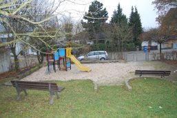 Ein Spielplatz mit Rutschturm in einer Sandfläche, zwei Holzbänken und einer Straße im Hintergrund