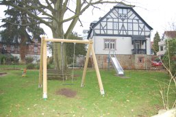 Ein Spielplatz mit Holzschaukel und Metallrutsche auf einer Wiese vor einem großen Fachwerkhaus