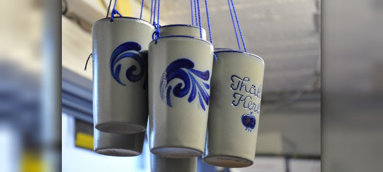 Eine Nahaufnahme von vier grau-blauen Keramikbechern, die an blauen Schnüren hängen. Die Becher sind mit blauen Wellenmustern verziert; auf einem ist der Schriftzug „Thäler Kerb“ und ein kleiner Apfel zu erkennen