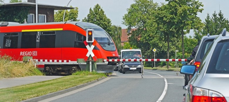 Ein roter Regionalzug passiert einen geschlossenen Bahnübergang mit rot-weiß gestreiften Schranken, vor denen mehrere Autos auf der Straße warten