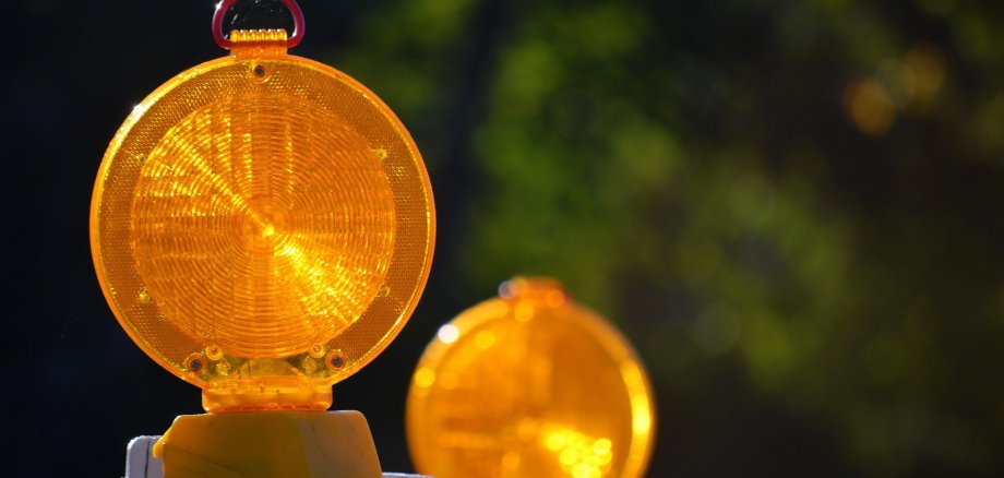 Zwei kreisförmige, gelbe Baustellenleuchten mit Prismenstruktur vor einem dunkelgrünen, unscharfen Hintergrund