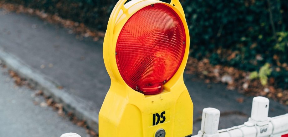 Gelbe Baustellen-Warnleuchte mit roter runder Linse, montiert auf einer rot-weiß gestreiften Absperrschranke an einem Gehweg