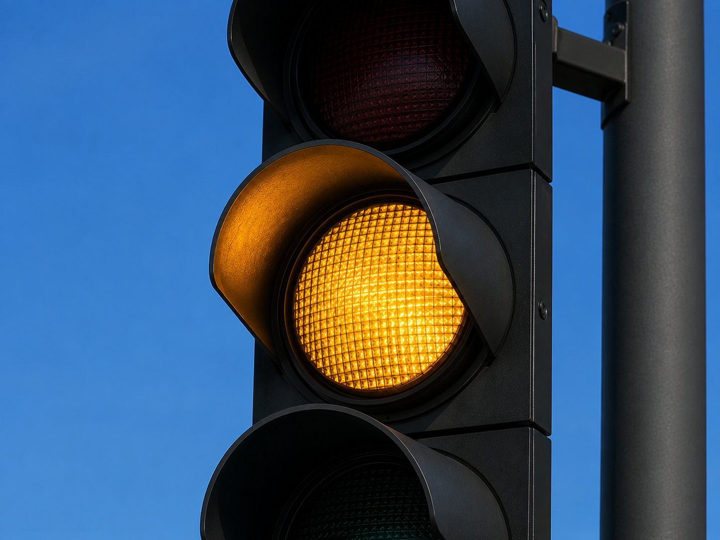 Nahaufnahme einer modernen schwarzen Straßenampel. Das mittlere, gelbe Licht leuchtet intensiv, während das obere rote und das untere grüne Licht ausgeschaltet sind. Im Hintergrund ist ein wolkenloser, blauer Himmel zu sehen