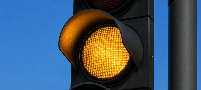 Ampel steht auf Gelb Nahaufnahme einer modernen schwarzen Straßenampel. Das mittlere, gelbe Licht leuchtet intensiv, während das obere rote und das untere grüne Licht ausgeschaltet sind. Im Hintergrund ist ein wolkenloser, blauer Himmel zu sehen