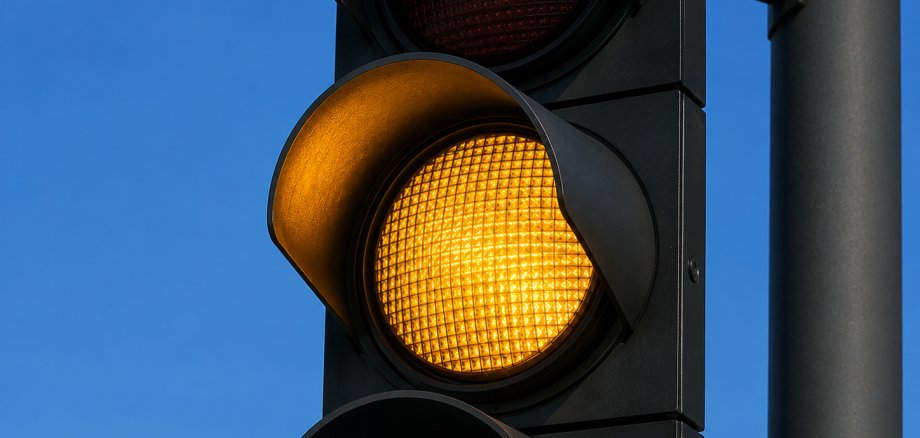 Ampel steht auf Gelb Nahaufnahme einer modernen schwarzen Straßenampel. Das mittlere, gelbe Licht leuchtet intensiv, während das obere rote und das untere grüne Licht ausgeschaltet sind. Im Hintergrund ist ein wolkenloser, blauer Himmel zu sehen