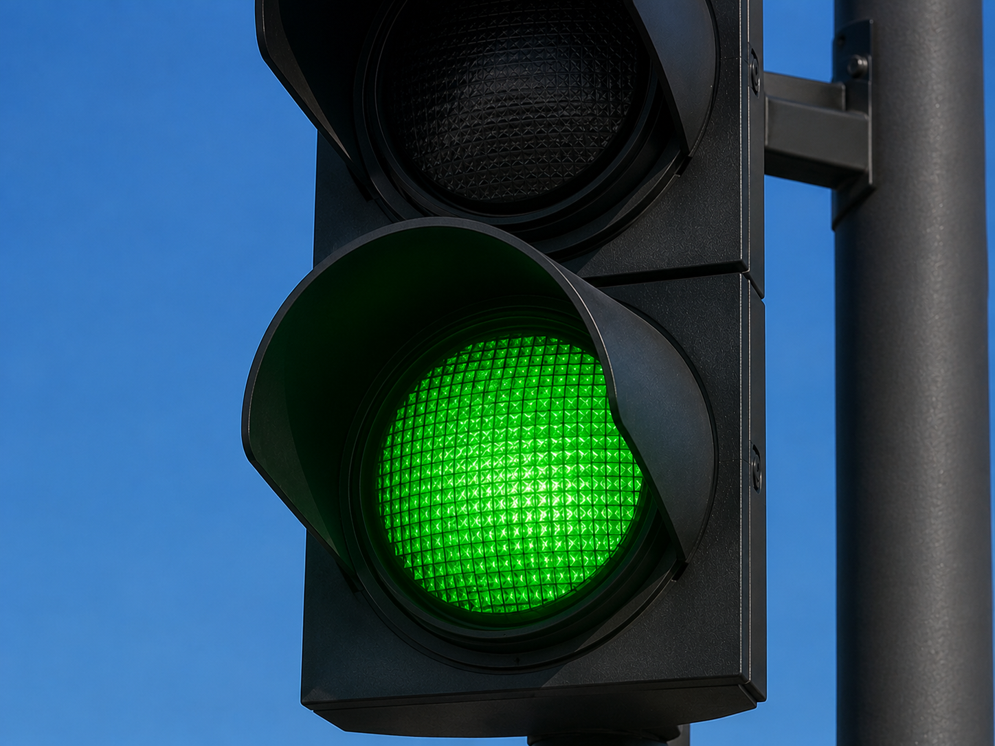 Ampel steht auf Grün Nahaufnahme einer modernen schwarzen Straßenampel. Das untere, grüne Licht leuchtet intensiv, während das mittlere gelbe und das obere rote Licht ausgeschaltet sind. Im Hintergrund ist ein wolkenloser, blauer Himmel zu sehen