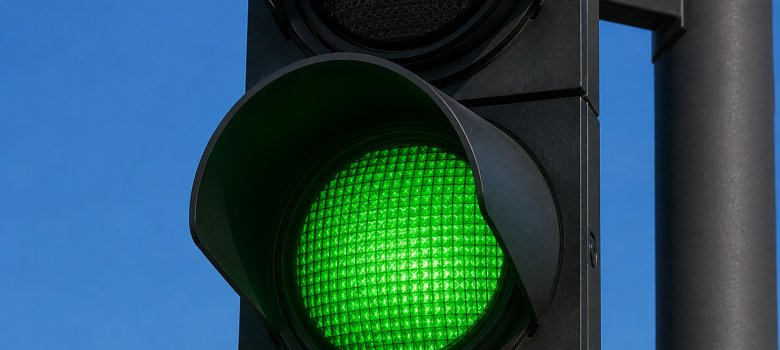 Ampel steht auf Grün Nahaufnahme einer modernen schwarzen Straßenampel. Das untere, grüne Licht leuchtet intensiv, während das mittlere gelbe und das obere rote Licht ausgeschaltet sind. Im Hintergrund ist ein wolkenloser, blauer Himmel zu sehen