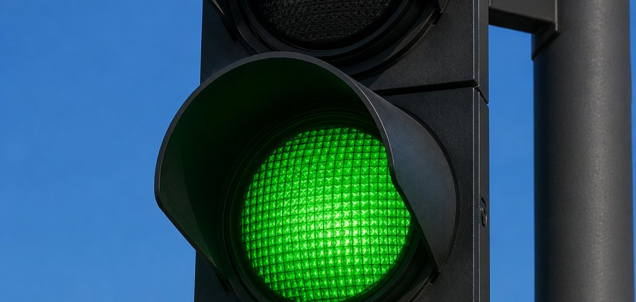 Nahaufnahme einer modernen schwarzen Straßenampel. Das untere, grüne Licht leuchtet intensiv, während das mittlere gelbe und das obere rote Licht ausgeschaltet sind. Im Hintergrund ist ein wolkenloser, blauer Himmel zu sehen