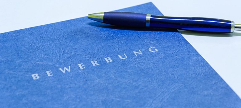Nahaufnahme einer strukturierten, dunkelblauen Klemmmappe mit silberner Prägung ‚BEWERBUNG‘, auf der ein farblich passender Kugelschreiber liegt