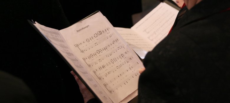 Rückansicht einer Person bei einem Chorkonzert, die eine Notenmappe in den Händen hält. Die Kamera fokussiert auf die Notenschrift und den Liedtitel ‚Bischofssegen‘