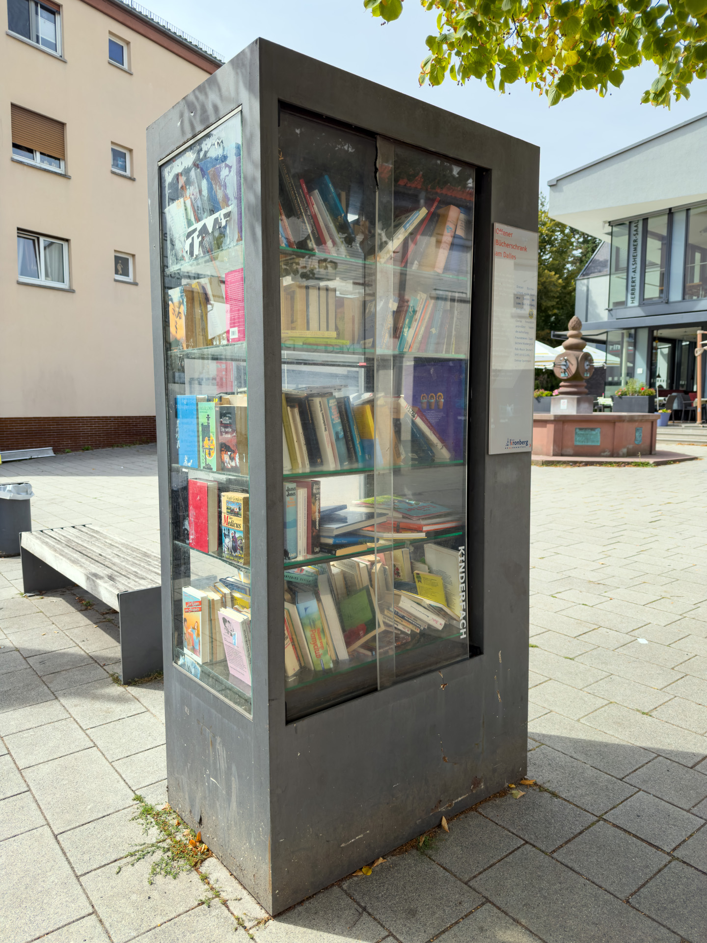 Ein moderner, grauer öffentlicher Bücherschrank aus Metall mit großen Glasscheiben auf einem gepflasterten Stadtplatz. Die Regale im Inneren sind dicht mit Büchern bestückt. Im Hintergrund sind eine Holzbank, ein Wohngebäude und ein Brunnen unter Bäumen zu sehen. Am Schrank befindet sich ein Hinweisschild mit dem Logo der Stadt Kronberg