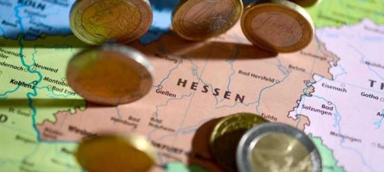 Nahaufnahme einer bunten Landkarte von Hessen; im Zentrum steht das Wort ‚HESSEN‘ in Großbuchstaben. Über die Karte sind scharf und unscharf fokussierte Euro-Münzen verteilt