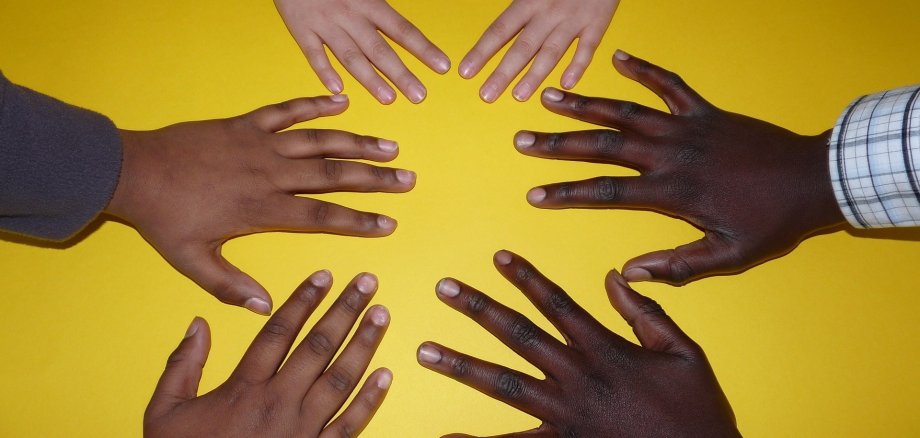 Draufsicht auf fünf Hände verschiedener Menschen, deren Fingerspitzen zur Mitte zeigen und so eine Sternform bilden. Der Hintergrund ist ein sattes, flächiges Gelb