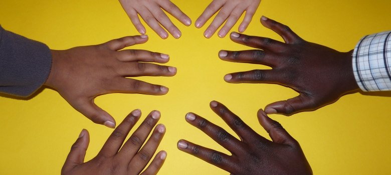Draufsicht auf fünf Hände verschiedener Menschen, deren Fingerspitzen zur Mitte zeigen und so eine Sternform bilden. Der Hintergrund ist ein sattes, flächiges Gelb