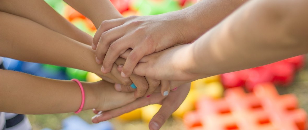 Mehrere Arme und Hände von Kindern liegen in der Bildmitte als Zeichen von Gemeinschaft übereinander gestapelt. Im unscharfen Hintergrund sind bunte Spielsteine aus Kunststoff auf einer Rasenfläche zu erkennen
